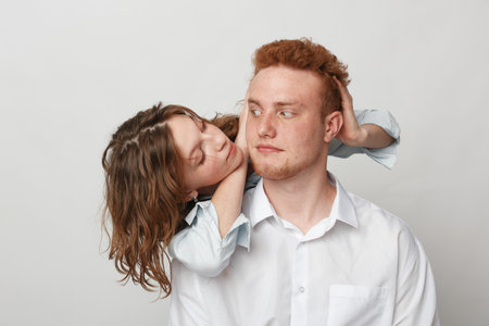 Young man with red hair and white shirt looks sideways while woman with curly hair hugs him from...