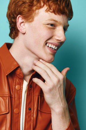 Young man with red hair smiling and touching his face wearing a brown leather jacket against a blue...