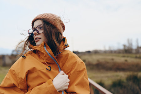 Young woman lifestyle outdoors wearing orange jacket and knitted hat looks away thoughtfully. Autumn...