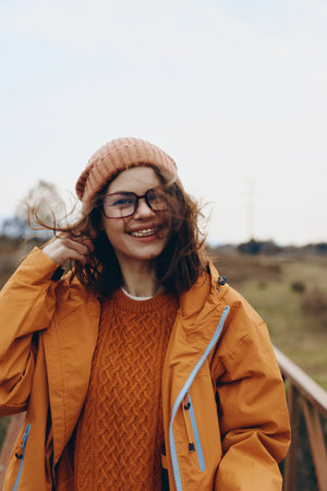 Smiling woman lifestyle outdoors wearing orange jacket and knit hat with glasses. Wind blows hair in...