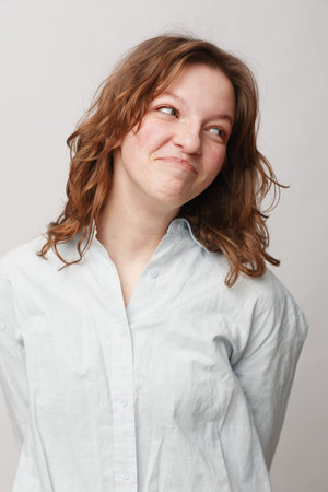 Young woman with curly hair wearing white shirt looks sideways with playful smile on light...