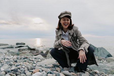 Happy young woman wearing a coat and hat laughs joyfully while sitting on a rocky beach during...