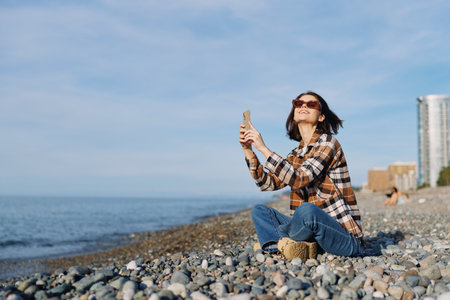 Woman sits on a pebbled beach by the sea, wearing a plaid shirt and jeans, taking a selfie with a...