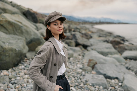 Young woman in stylish checkered coat and cap stands on rocky beach with calm sea and cloudy sky in background. Concept of fashion, autumn, and outdoor lifestyle portrait.の写真素材