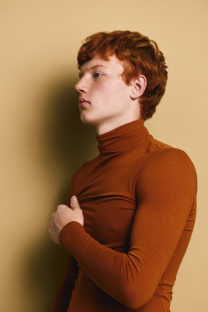 Young man with red hair wearing brown turtleneck standing sideways against beige background. Profile...