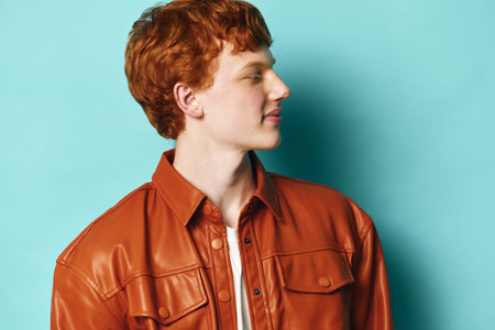 Young man with red hair wearing brown leather jacket and white shirt stands against blue background...