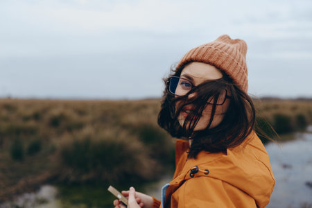 Young woman in orange jacket and brown hat holds smartphone outdoors. She looks back with hair...