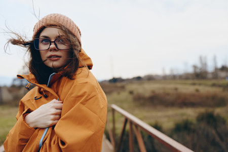 Young woman lifestyle outdoors wearing orange jacket and knitted hat stands on wooden bridge in...