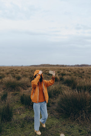 Woman lifestyle concept showing a female in casual orange jacket and jeans taking a selfie outdoors in a vast grassy field under a cloudy sky during daytime.の写真素材