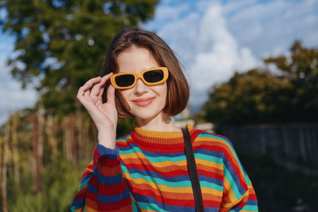 Woman, young woman, striped sweater, yellow sunglasses, smiling with short bob hair in a casual outfit outdoors; colorful knit, shoulder bag, sunlit urban park portrait for lifestyle travel.の写真素材