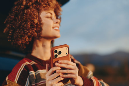 Smiling woman in rainbow sweater holds smartphone near car outdoors during golden hour, enjoying...