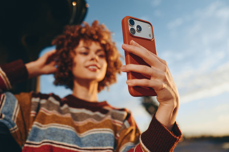 Young woman with curly hair wearing rainbow sweater takes selfie with smartphone outdoors in natural...