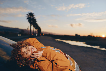 Smiling woman with curly hair wearing a rainbow sweater and orange jacket talking on smartphone near...
