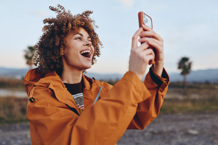 A happy woman with curly hair wearing an orange jacket smiles broadly while holding a smartphone...