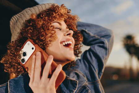 A happy woman with curly hair wearing a beige beanie and denim jacket smiles while talking on a...