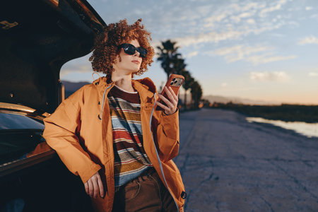 A woman with curly hair wearing sunglasses and a rainbow sweater smiles while leaning on a car and...