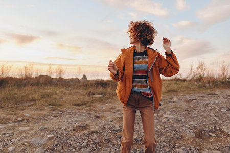 Woman with curly hair wearing orange jacket stands outdoors on rocky ground during sunset smiling...
