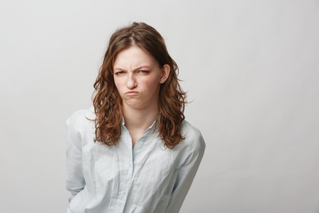 Young woman with curly hair wearing white shirt shows displeased facial expression, frowning and...