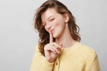 Young woman in yellow sweater makes shushing gesture with finger on lips, smiling slightly. Studio...