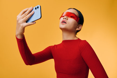 Young woman wearing red translucent sunglasses and red sweater takes a selfie with smartphone...