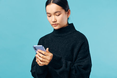 Young woman with dark hair wearing black sweater, looking down at smartphone with focused expression...