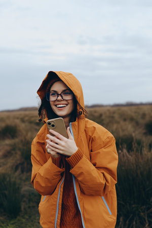 Smiling woman in orange jacket and glasses holds smartphone outdoors in natural landscape. Lifestyle...