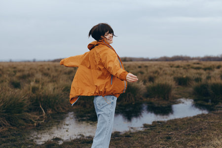 Young woman lifestyle outdoors in casual orange jacket and jeans spins happily near wetland under...