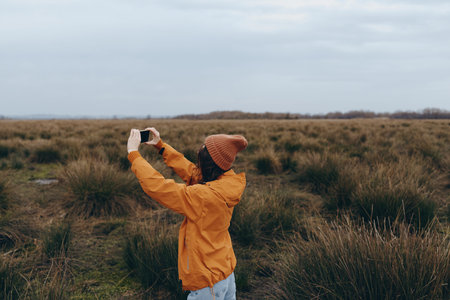 Woman lifestyle outdoors in autumn field wearing orange jacket and hat takes photo with smartphone....