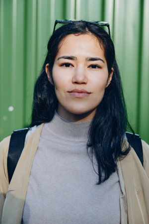 Woman with glasses on head wearing gray turtleneck and beige jacket stands before a vivid green...