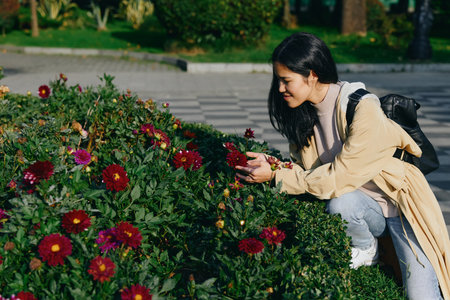 Woman crouching beside blooming flowers in a garden, reaching to pick petals, carrying a backpack...