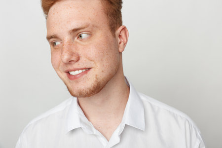 Young redhead man with freckles smiles looking sideways wearing white shirt on light background....