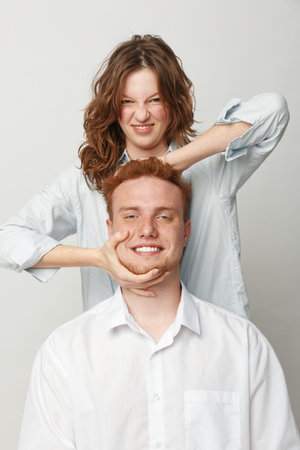 Playful young woman with curly hair teasing smiling redhead man in white shirts against plain...