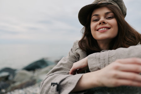 Smiling young woman in casual clothes and hat leans on a rock at the beach. Natural light highlights her relaxed mood and outdoor lifestyle on a cloudy day by the sea.の写真素材