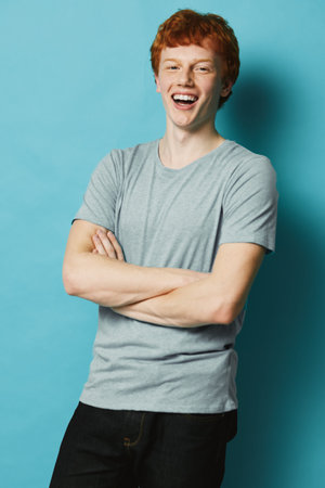 Young man with red hair smiling and laughing with arms crossed wearing gray t shirt and black pants...