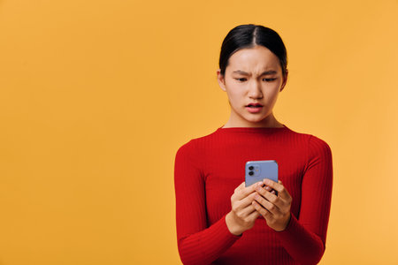 Young woman in red sweater looking confused at smartphone screen with yellow background. Female...