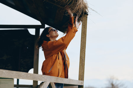 Woman lifestyle concept with a young woman wearing an orange jacket reaching up to adjust a wooden roof on a sunny day outdoors. Casual fashion and nature background.の写真素材