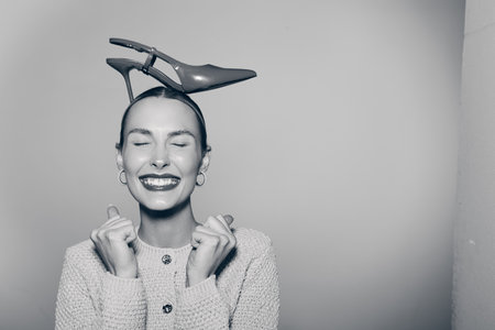 Happy caucasian woman with light skin smiles with closed eyes wearing high heel shoe on head, posing...
