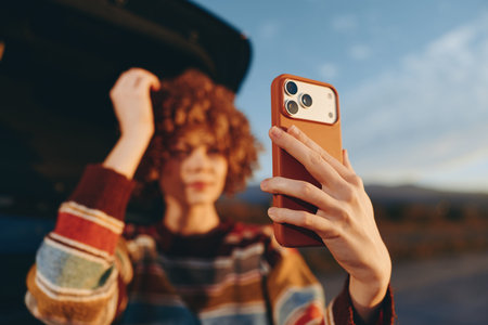 Smiling woman with curly hair wearing a rainbow sweater takes a selfie with a smartphone outdoors in...