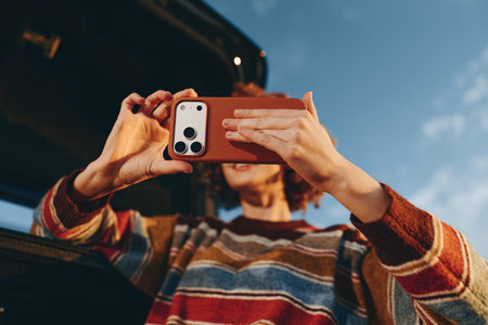 Woman wearing rainbow sweater smiling while taking a selfie with smartphone near car under clear...