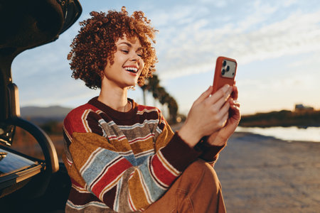 Happy woman with curly hair wearing rainbow sweater smiles while sitting near car outdoors using...