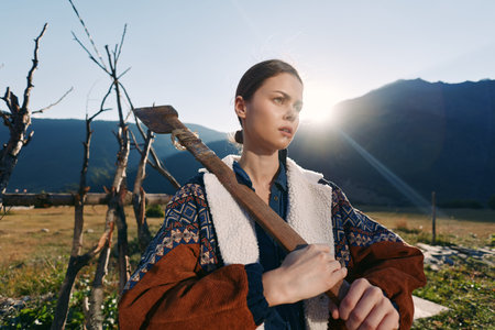 Woman with axe on shoulder on rural farm, portrait at sunrise in mountains. Outdoor worker in warm...