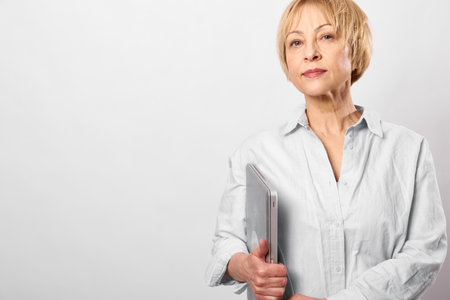 Confident mature woman with short blonde hair in white shirt holding laptop, standing against light...