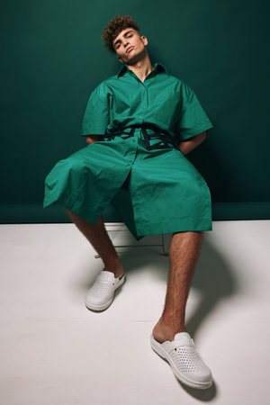 Young man wearing green oversized shirt and shorts sitting on bench with relaxed pose against dark...