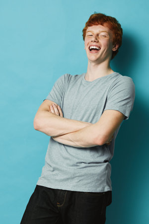 Young man with red hair wearing gray t-shirt laughing with arms crossed standing against blue...