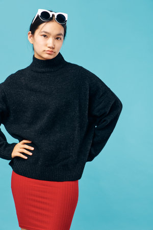 Young woman posing in black sweater and red skirt against blue background with white sunglasses on...