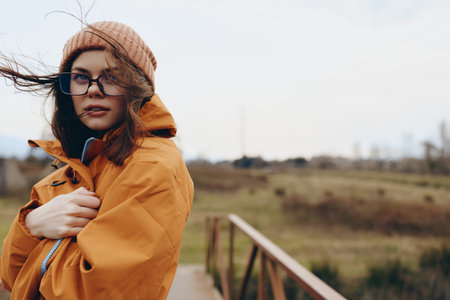 Young woman in orange jacket and beanie stands outdoors on a wooden bridge in windy weather....