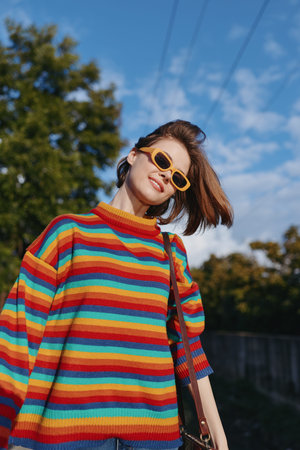 Young woman in rainbow sweater and yellow sunglasses smiling outdoors under blue sky. Early twenties...