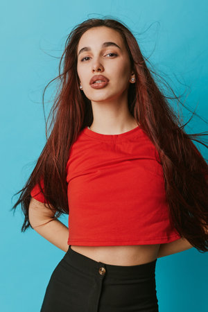 Young woman with long hair wearing red shirt and black pants posing confidently against blue...