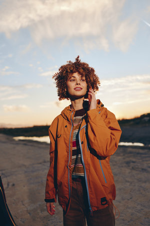 A smiling woman wearing a sweater and orange jacket talks on a smartphone outdoors during sunset....