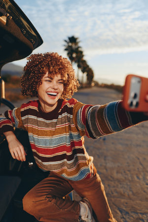 Cheerful woman with curly hair wearing sweater taking selfie with smartphone near car outdoors, smiling brightly, casual lifestyle, sunny day, palm trees in backgroundの写真素材